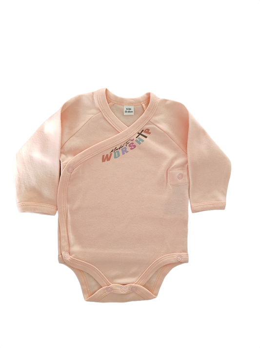 A baby long-sleeve wrap bodysuit in a soft peach-pink color, featuring a colorful 'Worship' graphic and a small cross on the chest.