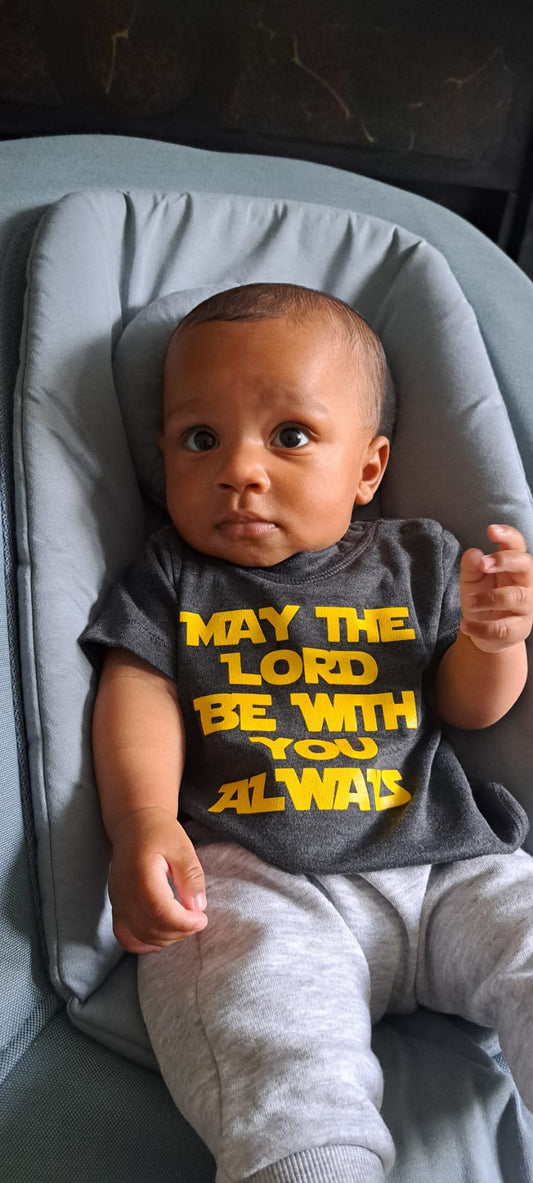 A baby wearing a grey bodysuit with the text 'MAY THE LORD BE WITH YOU ALWAYS' printed on the front.